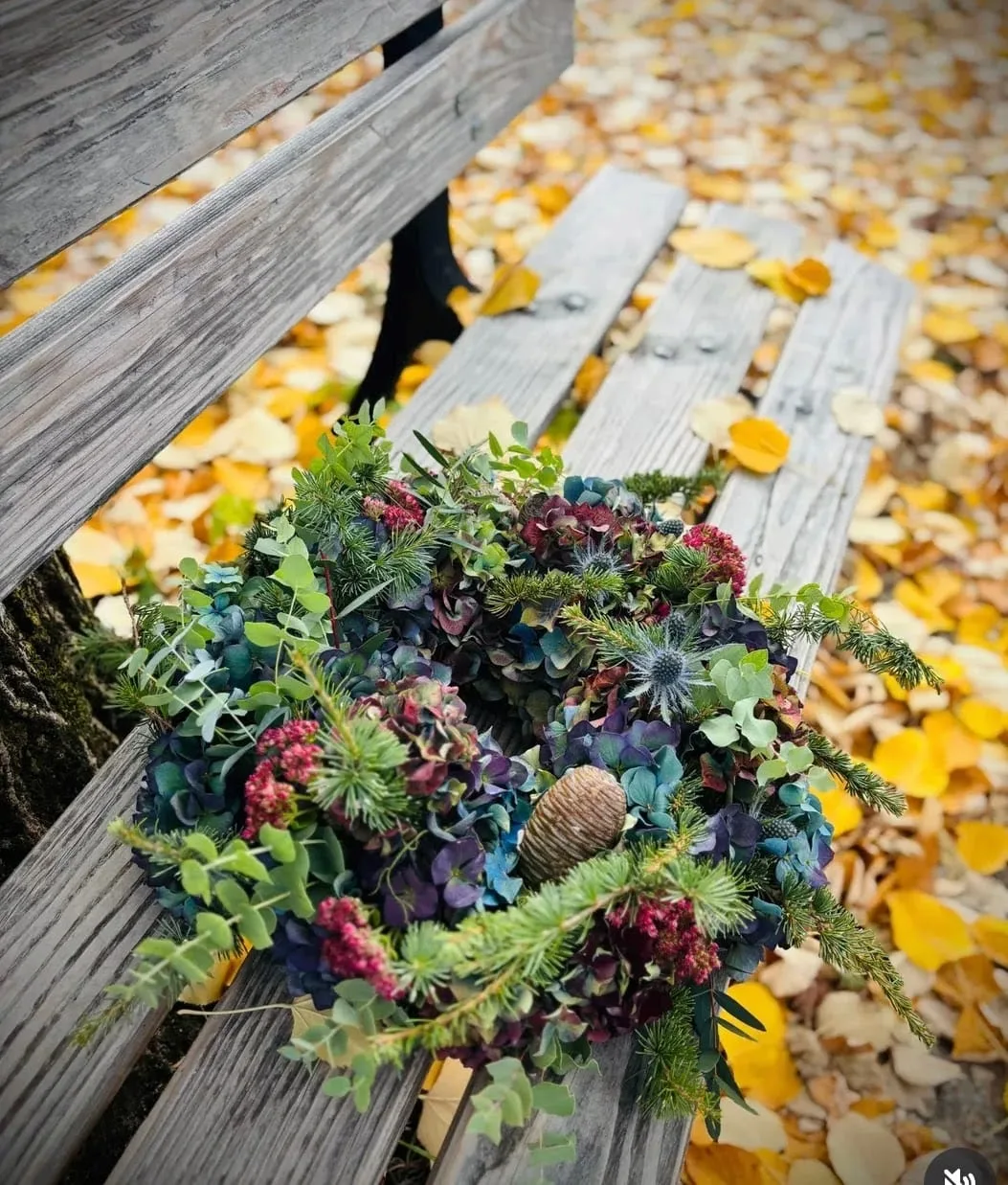 Kranz aus Hortensien, Eukalyptus, Disteln und Zapfen auf einer Holzbank im Herbstlaub