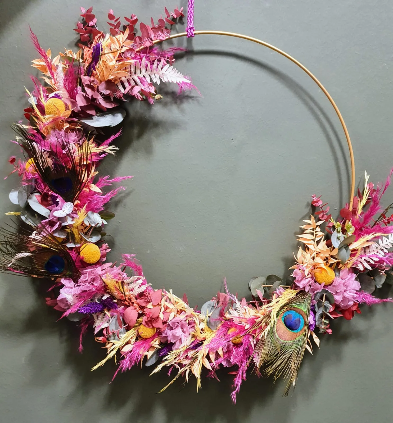 Großer Trockenblumen-Loop in Pink und Gelb mit Pfauenfeder an einer grünen Wand