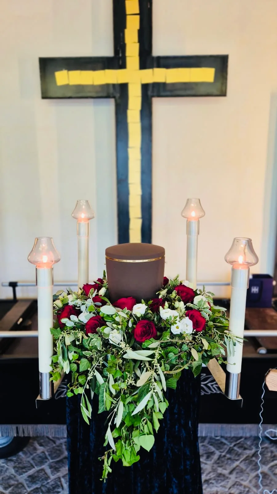 Urne mit Schmuck aus roten Rosen und Grün, daneben Kerzen und ein Kreuz