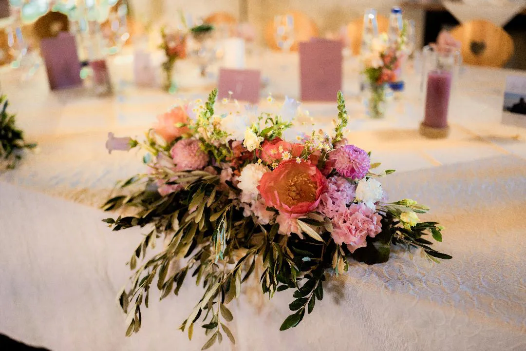 Niedriges Gesteck aus Rosen, Pfingstrosen und Olivenzweigen auf einer weiß gedeckten Tafel