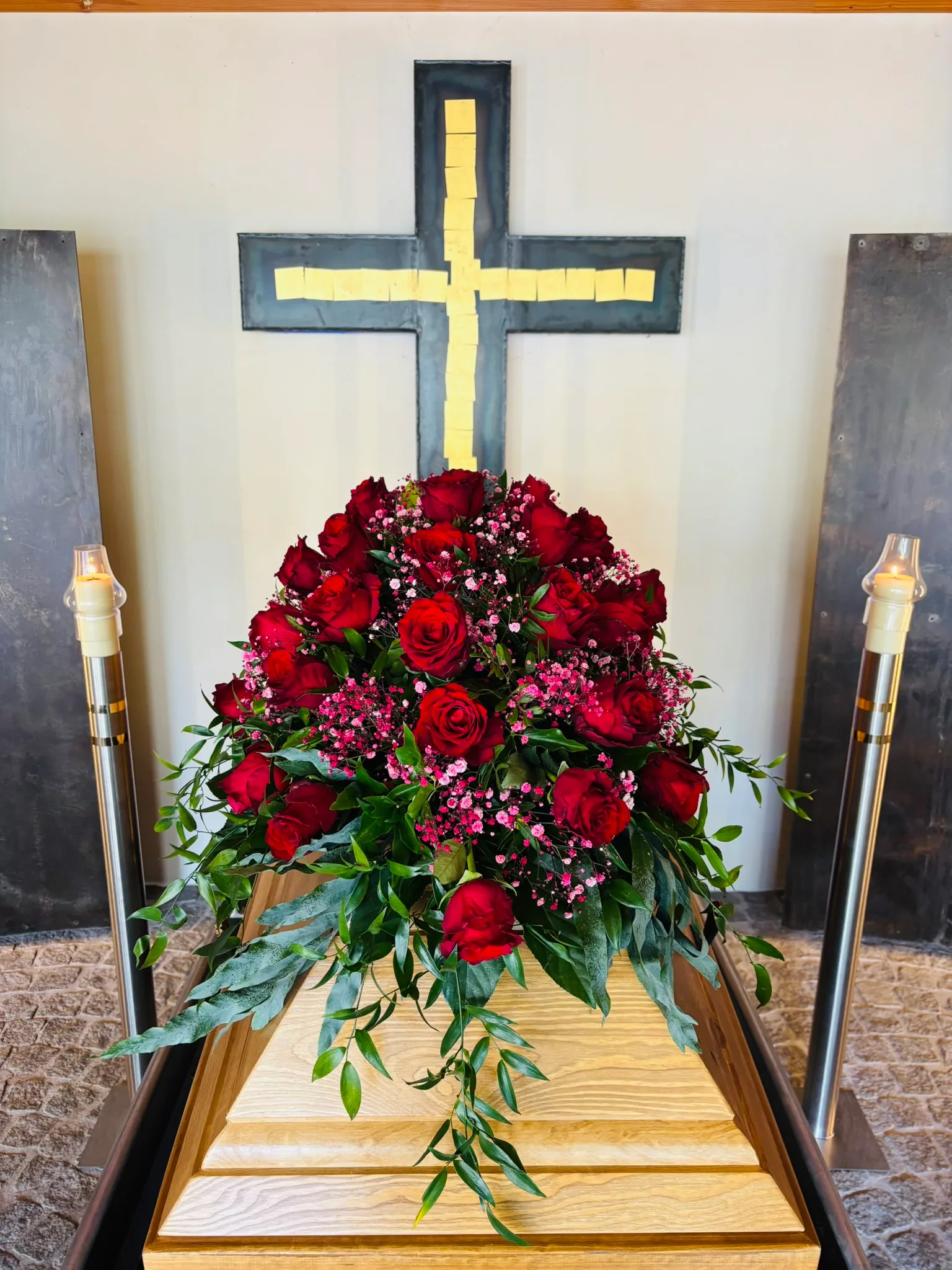 Sargschmuck aus roten Rosen auf einem Holzsarg, dahinter ein Kreuz