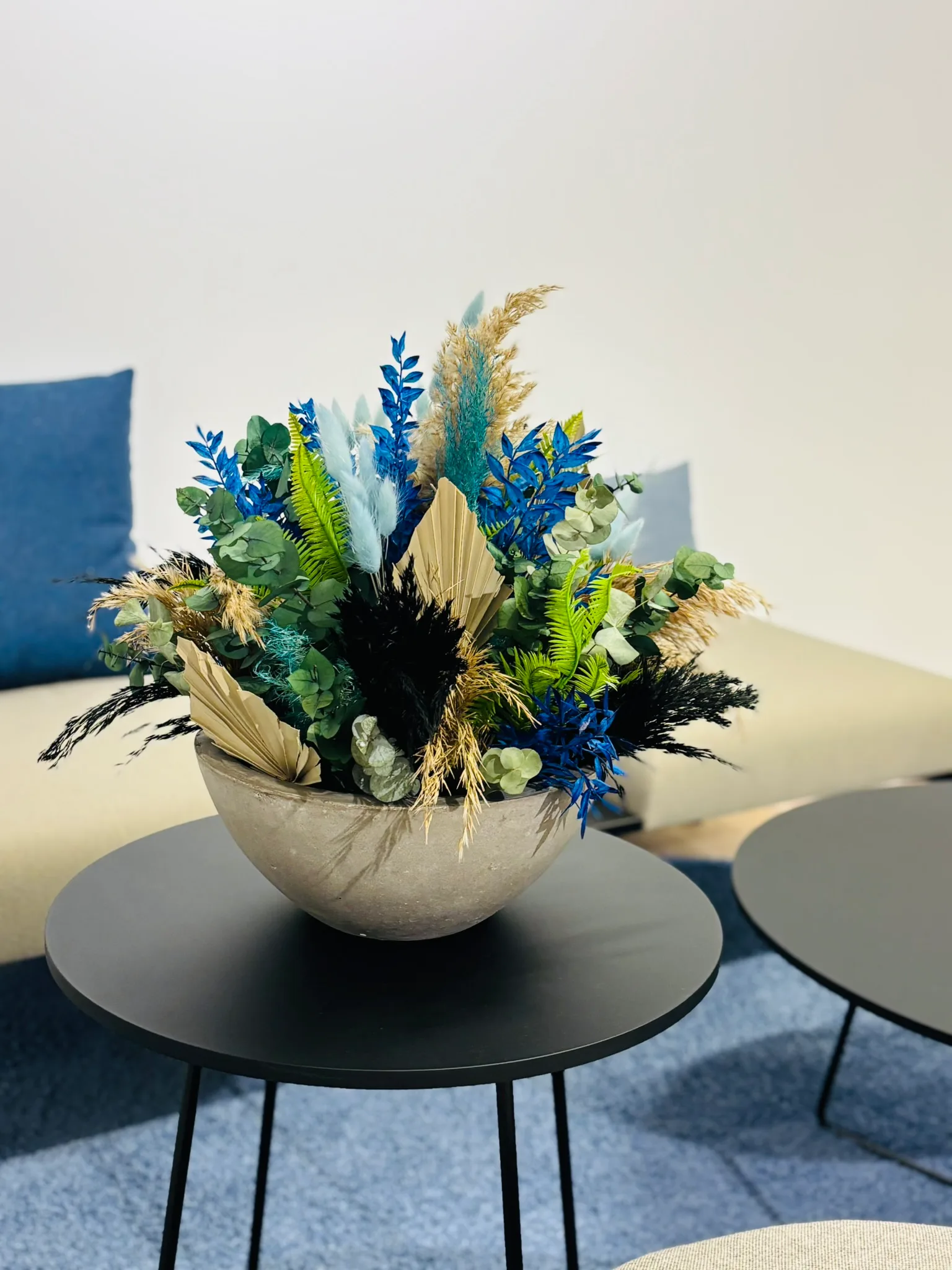 Gesteck aus Trockenblumen in Blau, Grün und Creme in einer Schale auf einem Loungetisch
