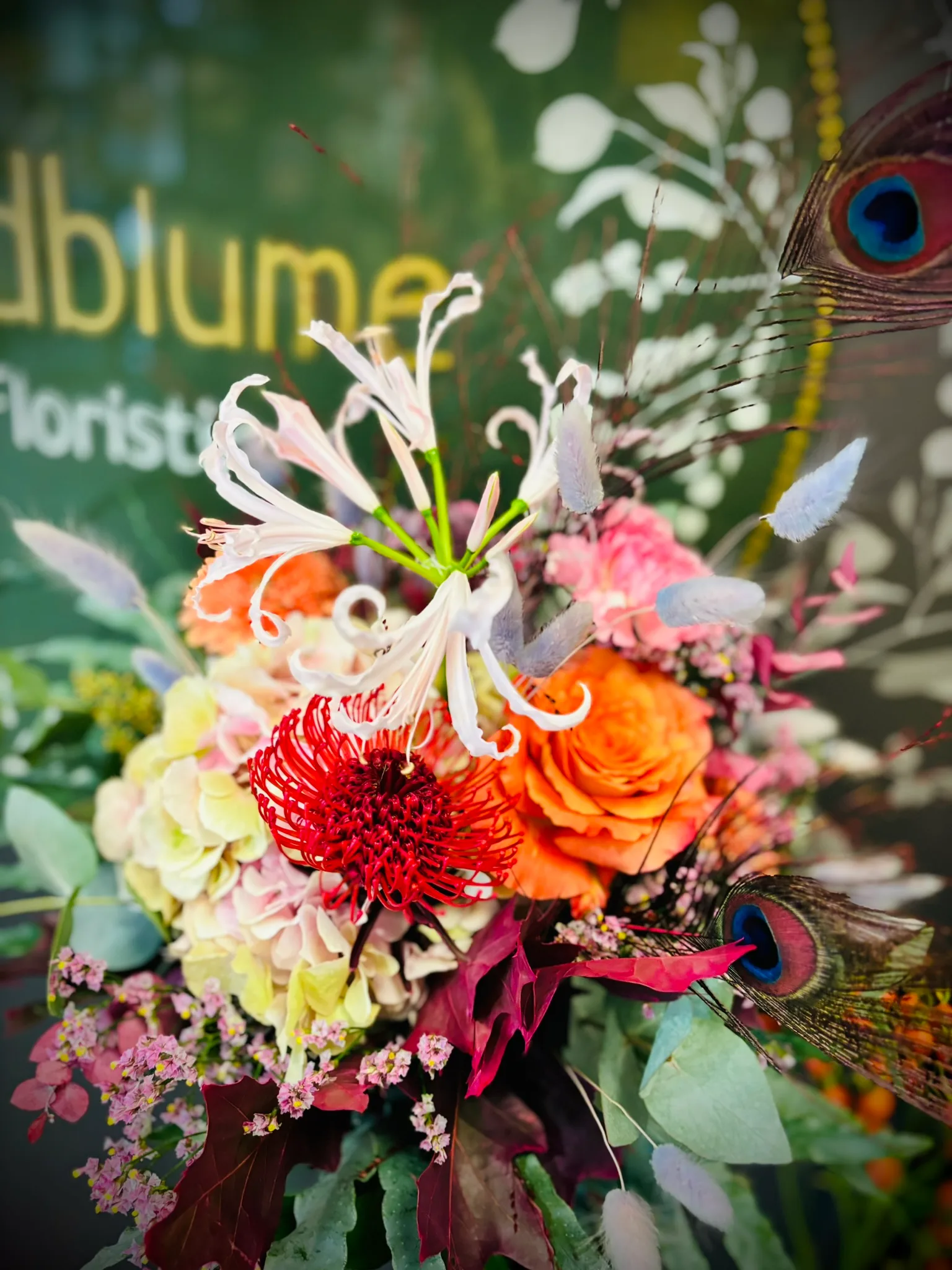 Strauß mit Nerine, Protea, orangefarbener Rose und Pfauenfeder vor dem Wildblume-Schild